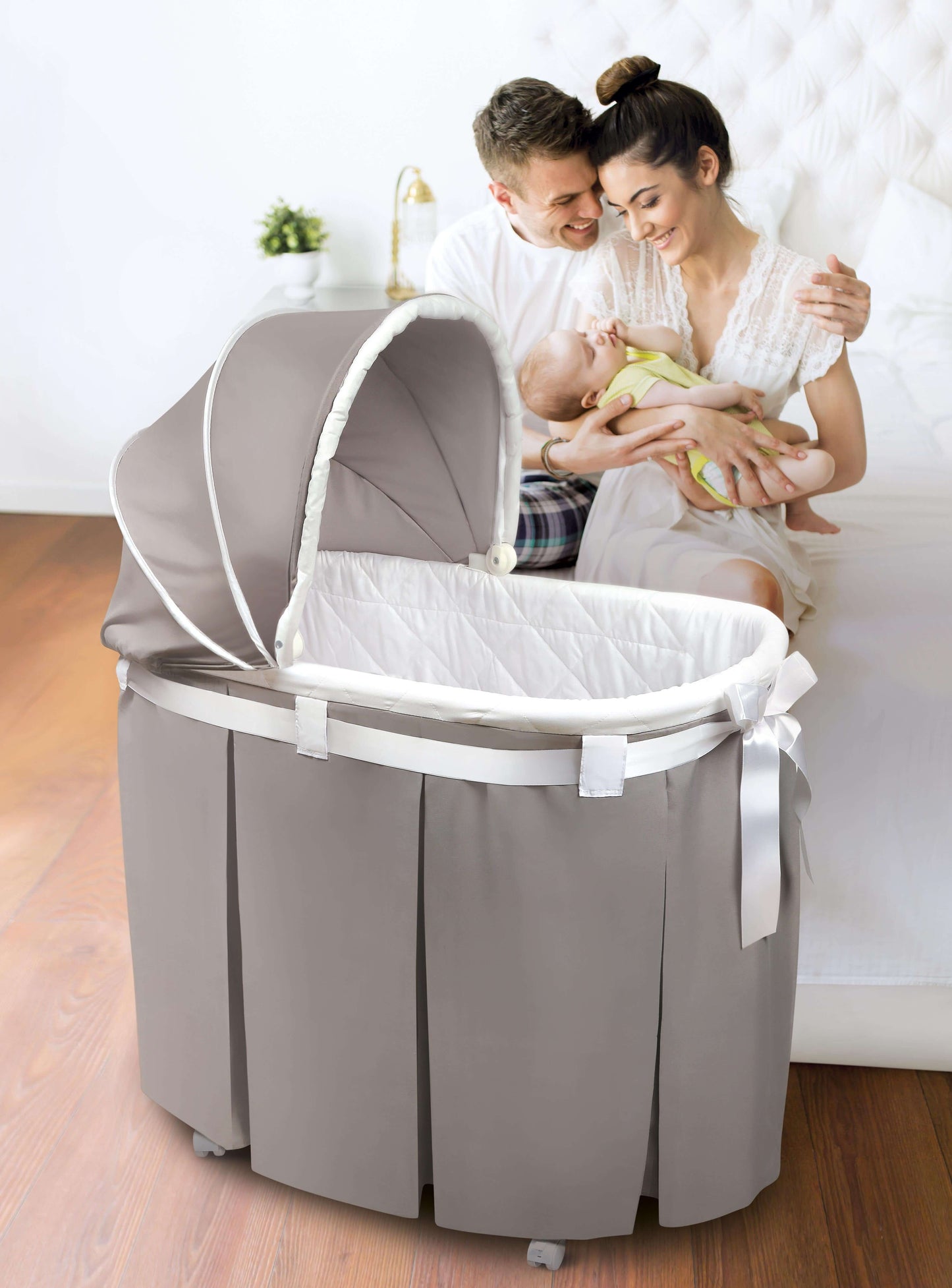 Wishes Oval Bassinet - Lifestyle Image of husband and Wife Holding Baby Besides Wishes Oval Bassinet Gray Bedding