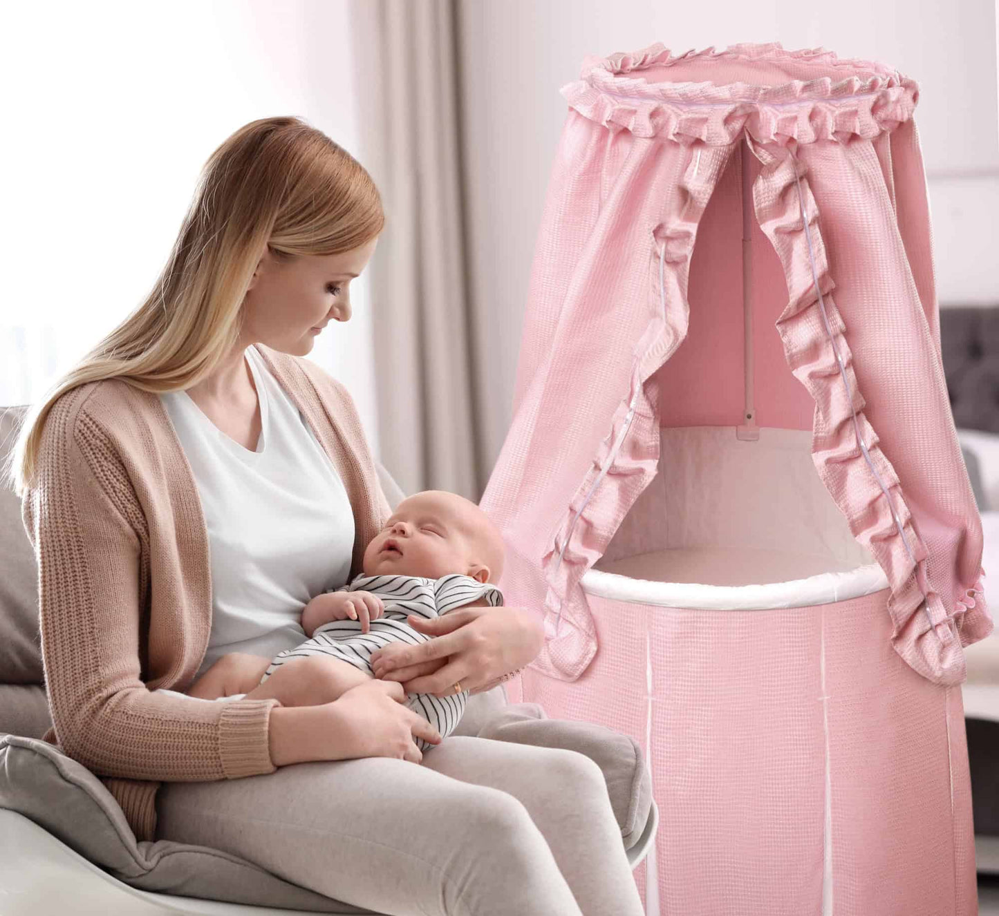 Empress Round Baby Bassinet with Canopy - Lifestyle Image of Mother Holding Baby, Sitting Beside Empress Round Baby Bassinet with Canopy Pink