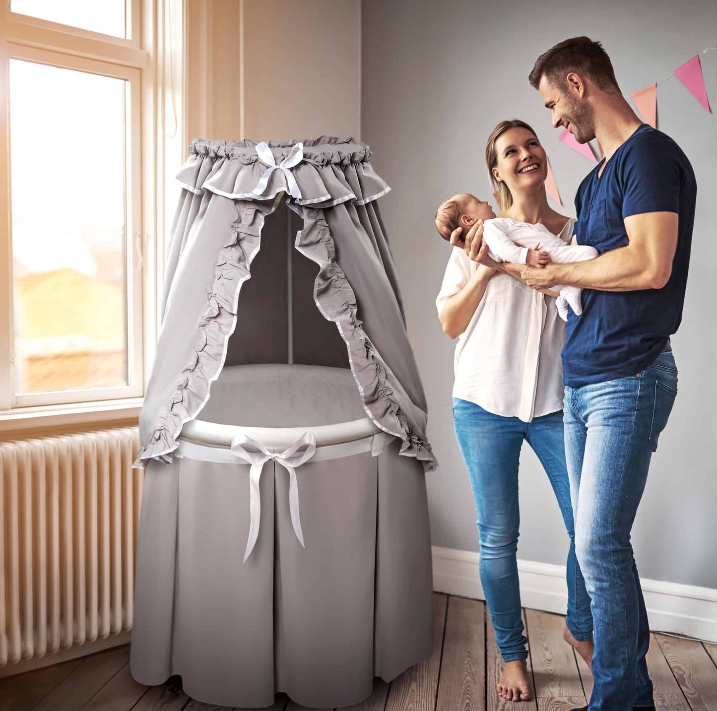 Empress Round Baby Bassinet with Canopy - Lifestyle Image of Man and Woman Holding Baby Besides Empress Round Baby Bassinet with Canopy Multi Tone Gray