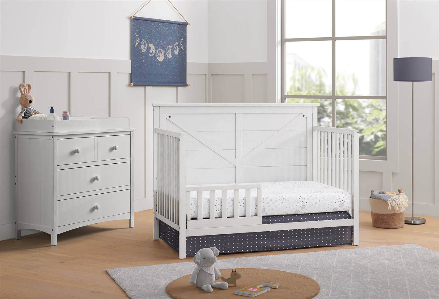 Montauk 4-in-1 Convertible Crib - Lifestyle Image Inclined Side View of Montauk 4-in-1 Convertible Crib Barn White with Guard Rail and 3 Drawer Dresser