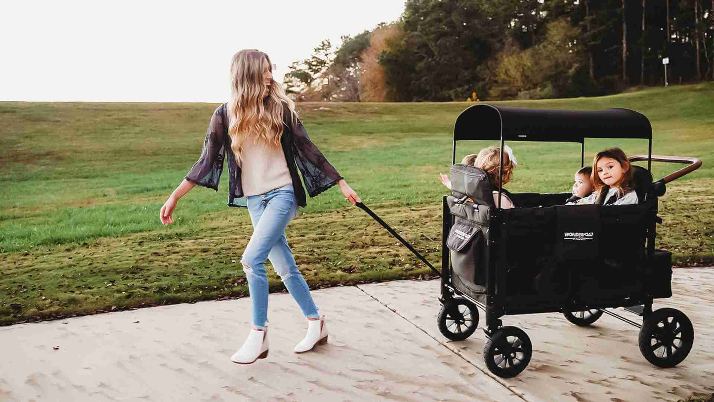 W4 Luxe Quad Stroller Wagon - Lifestyle Image Inclined View of Toddler's on W4 Luxe Quad Stroller Wagon With Mother pulling the Wagon