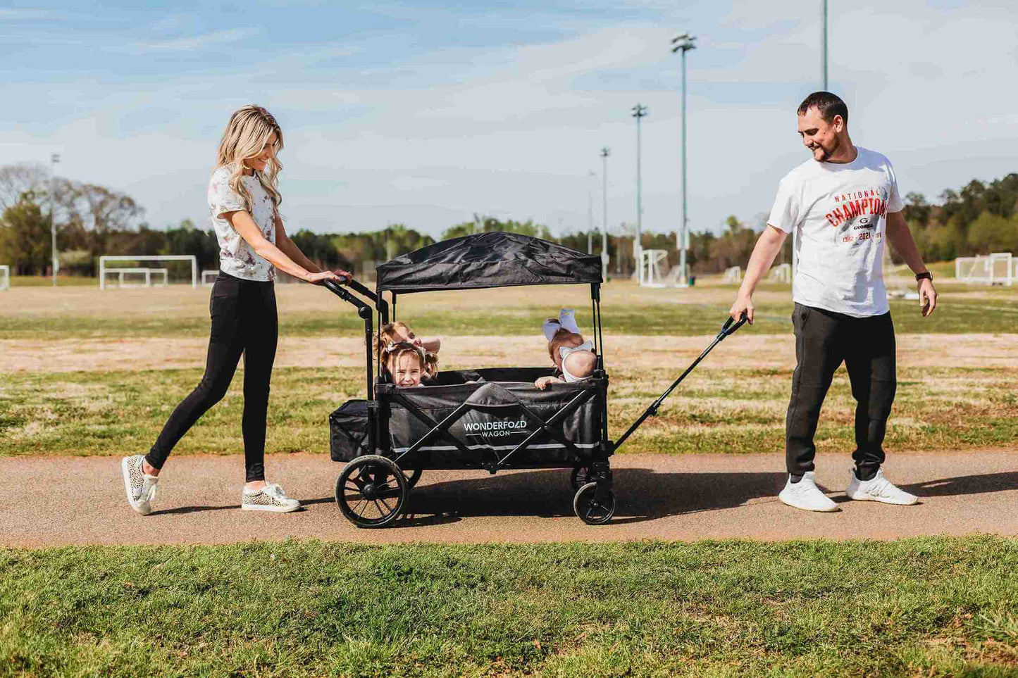 X4 Push + Pull Quad Stroller Wagon - Lifestyle Image Side View of Mother Holding Push Handle and Father Holding Pull Handle of X4 Push + Pull Quad Stroller Wagon Black With Kids Inside
